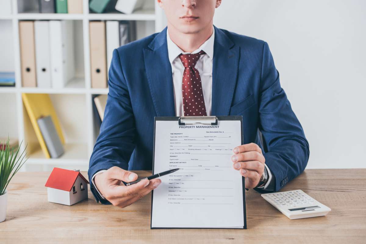 Cropped view of businessman holding clipboard with property management agreement near house model and calculator