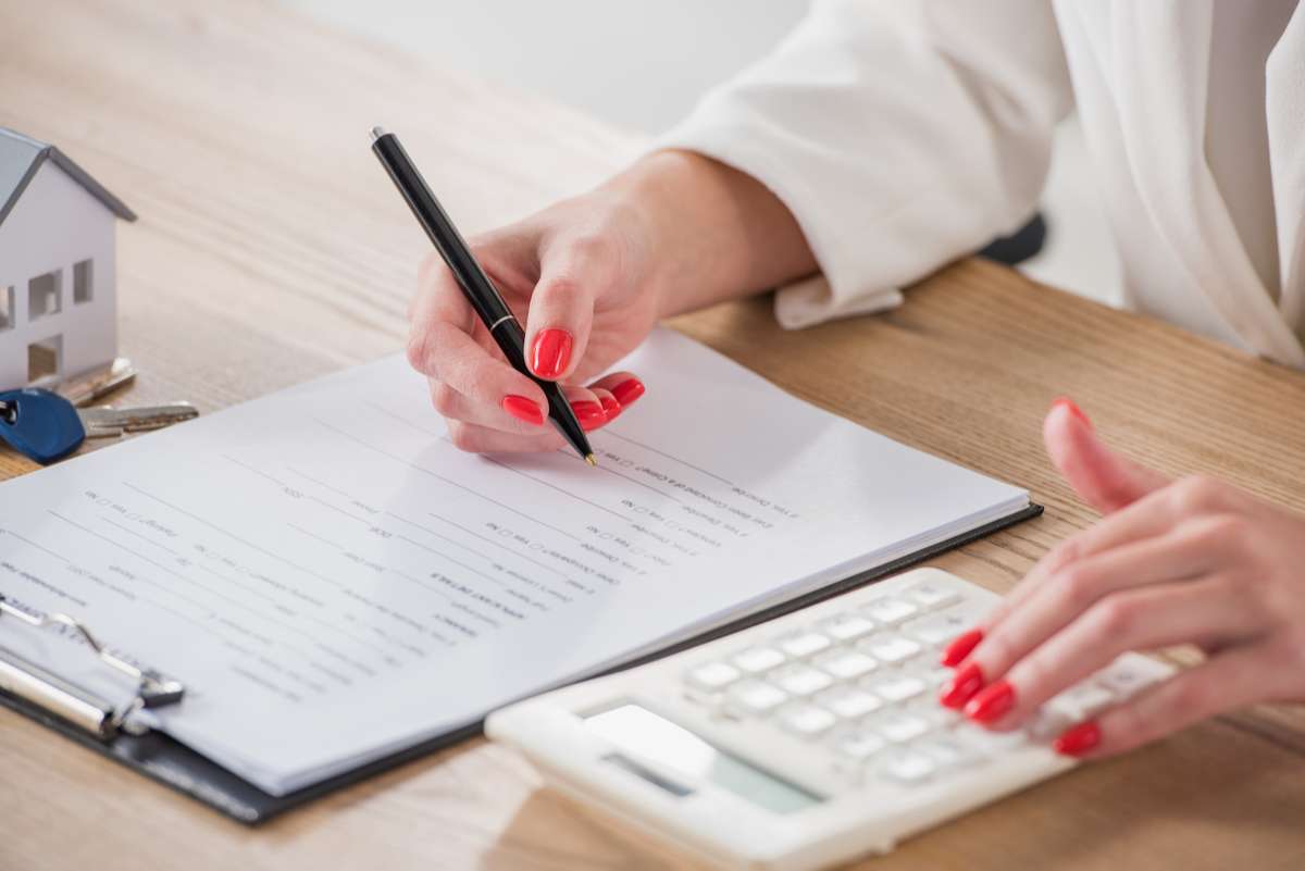 Cropped view of businesswoman using calculator and writing in contract near house model and keys