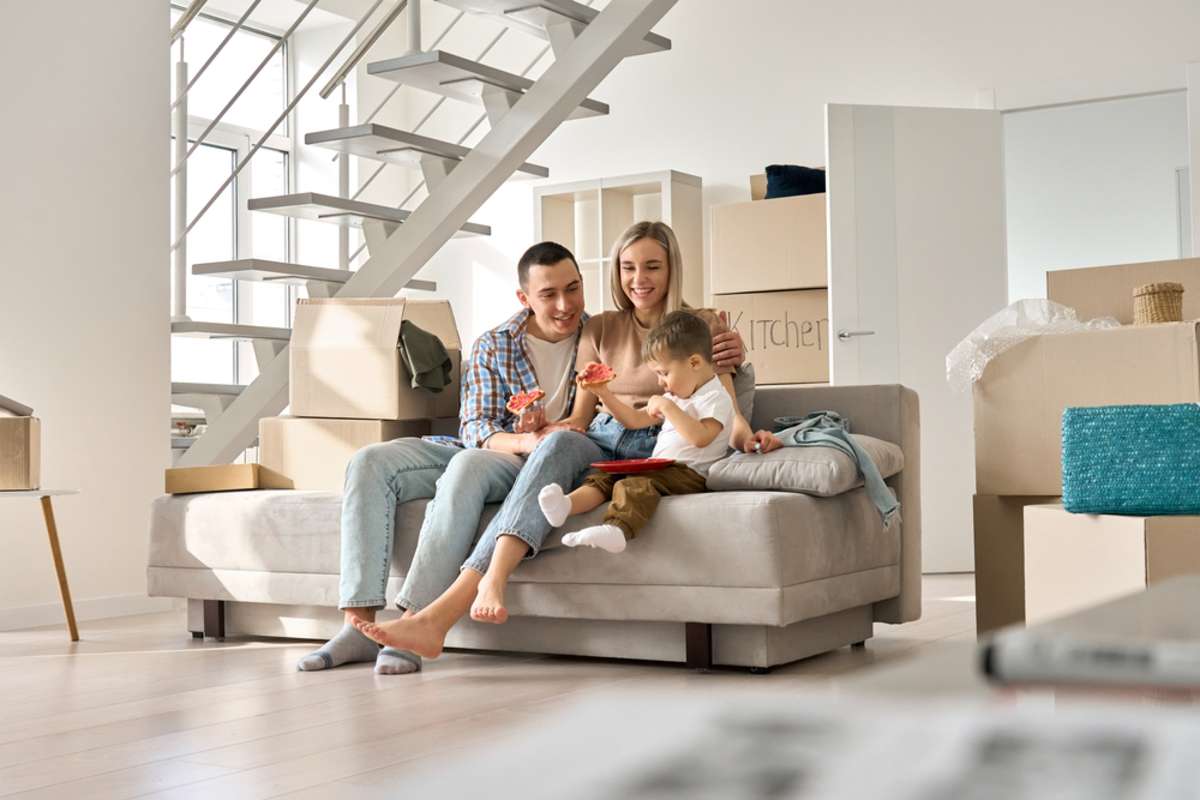 Happy family couple new home owners with kid child son relaxing on couch eating snack unpacking boxes on moving day in living room