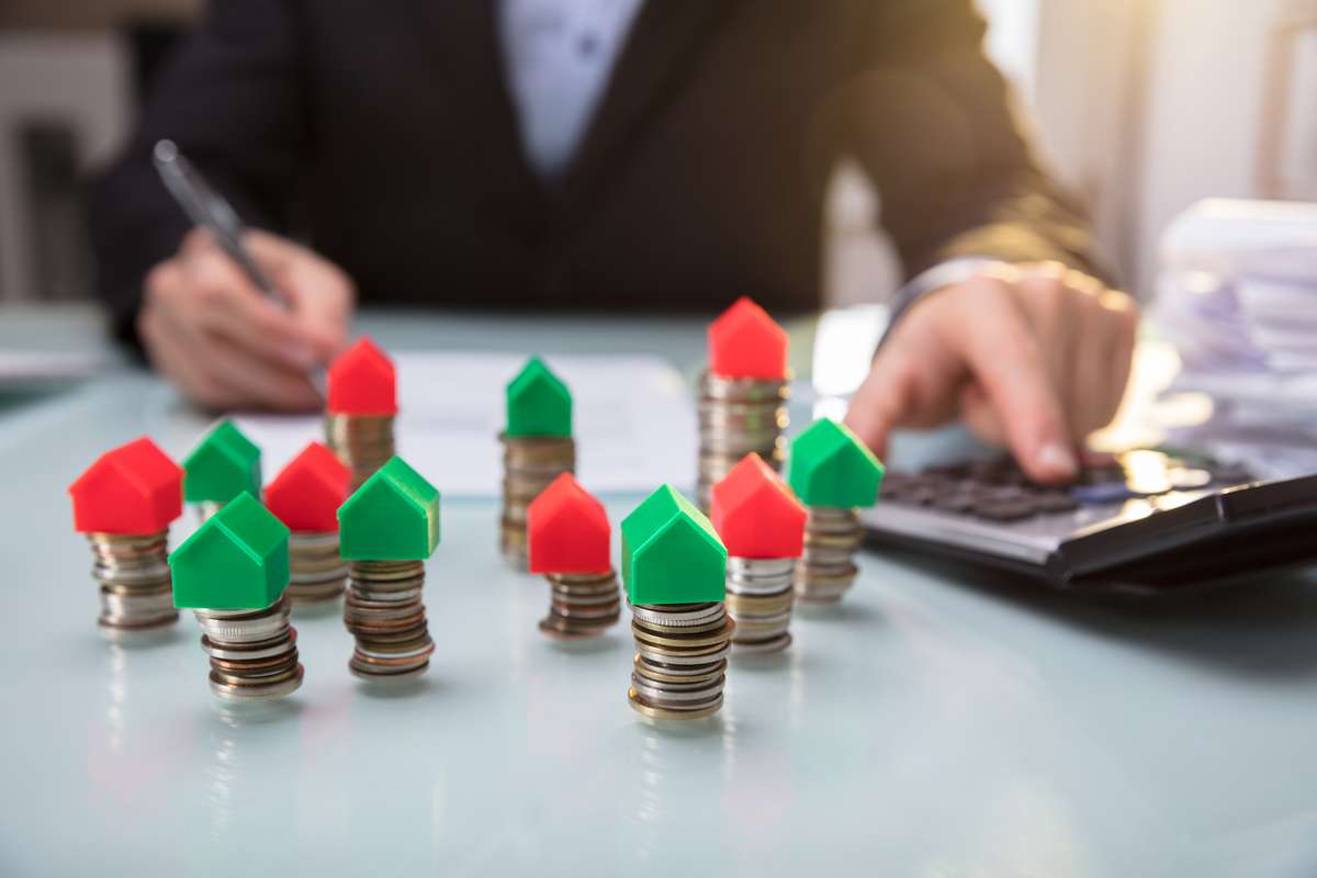 Close-up Of Green And Red House Model On Stacked Coins Besides Businesspersons Hand Using Calculator (R) (S)